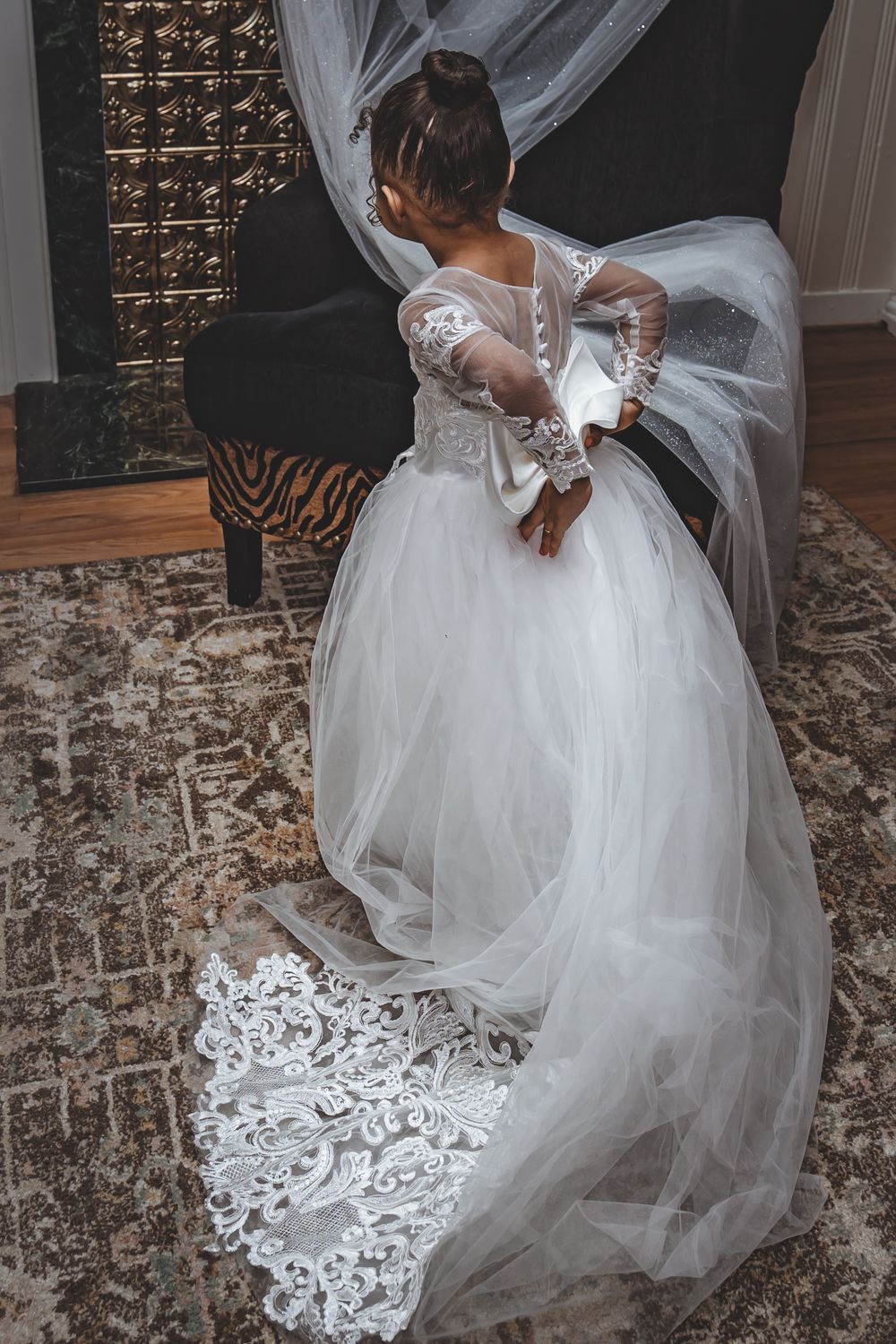 A bride in a white lace wedding dress with long train and veil stands next to an ornate fireplace.