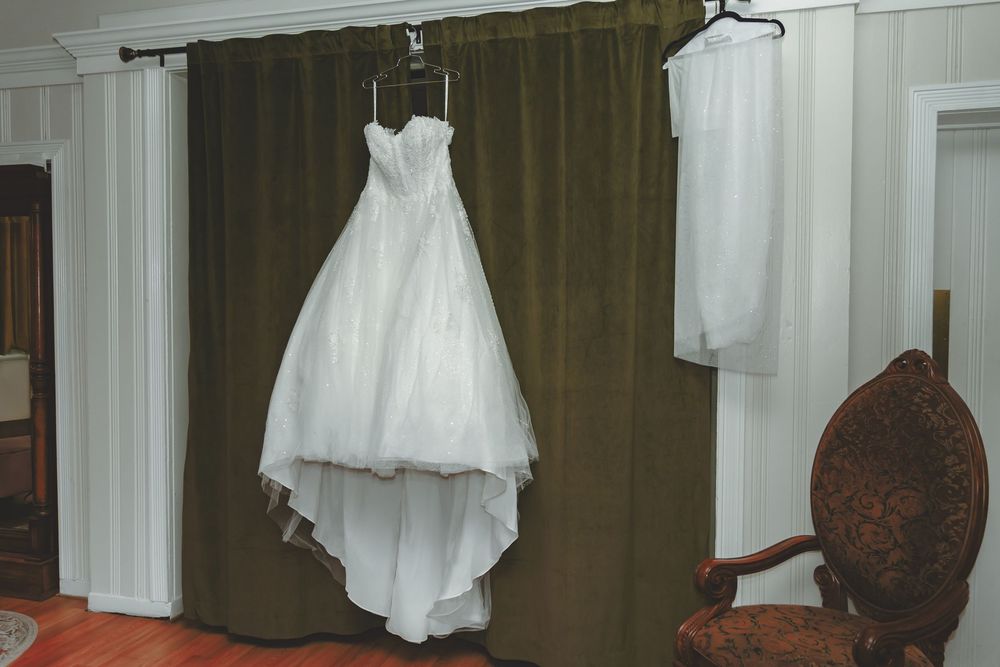 A white wedding dress with flowy tulle skirt hangs from a hanger against brown curtains in a fitting room.