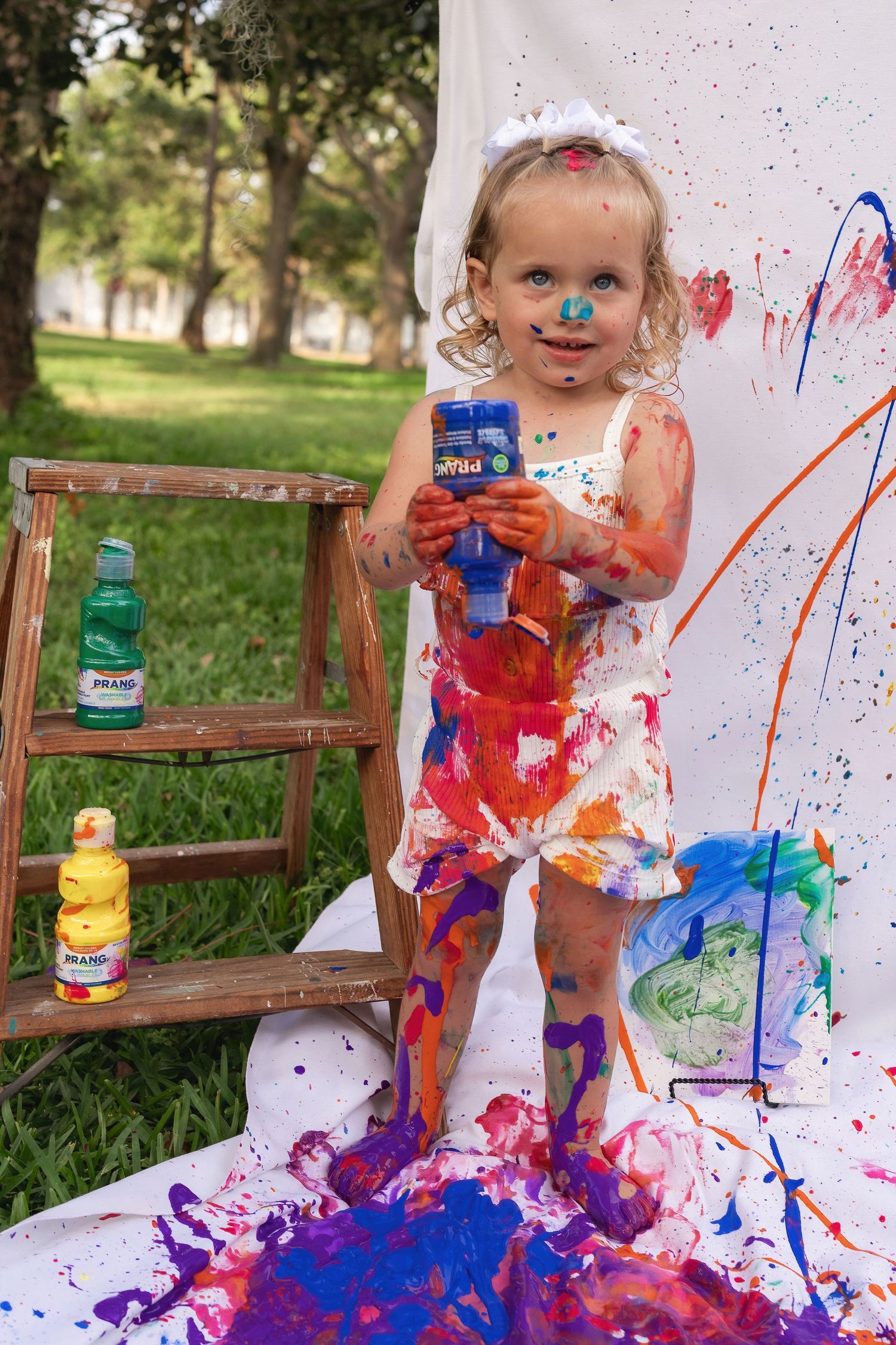 Small child covered in colorful paint splatters holds blue paint bottle during messy outdoor art session.