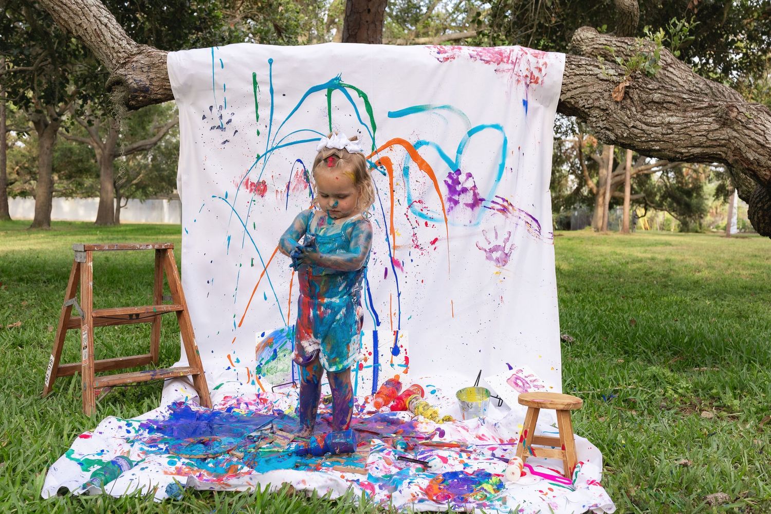 Child in blue clothes making abstract art with paint on white backdrop outdoors.