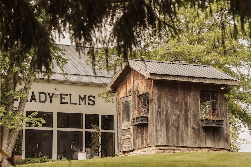 Rustic wooden barn building with 'SHADY ELMS' signage visible through overhanging tree branches in a rural setting.