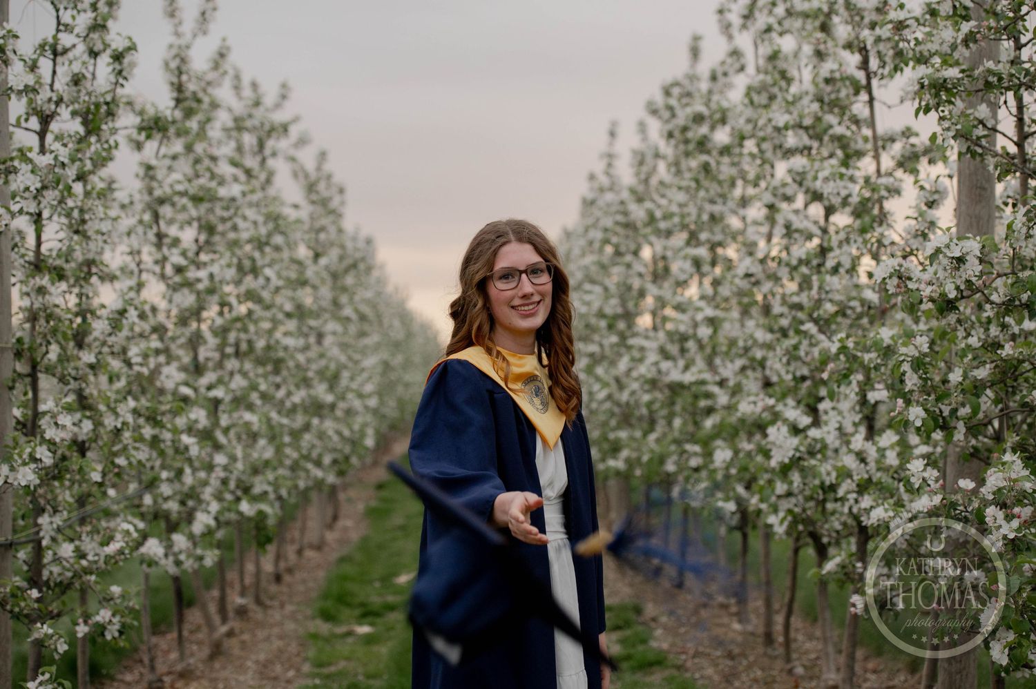 A person stands among blooming spring orchard trees at dusk wearing a navy blue graduation gown.