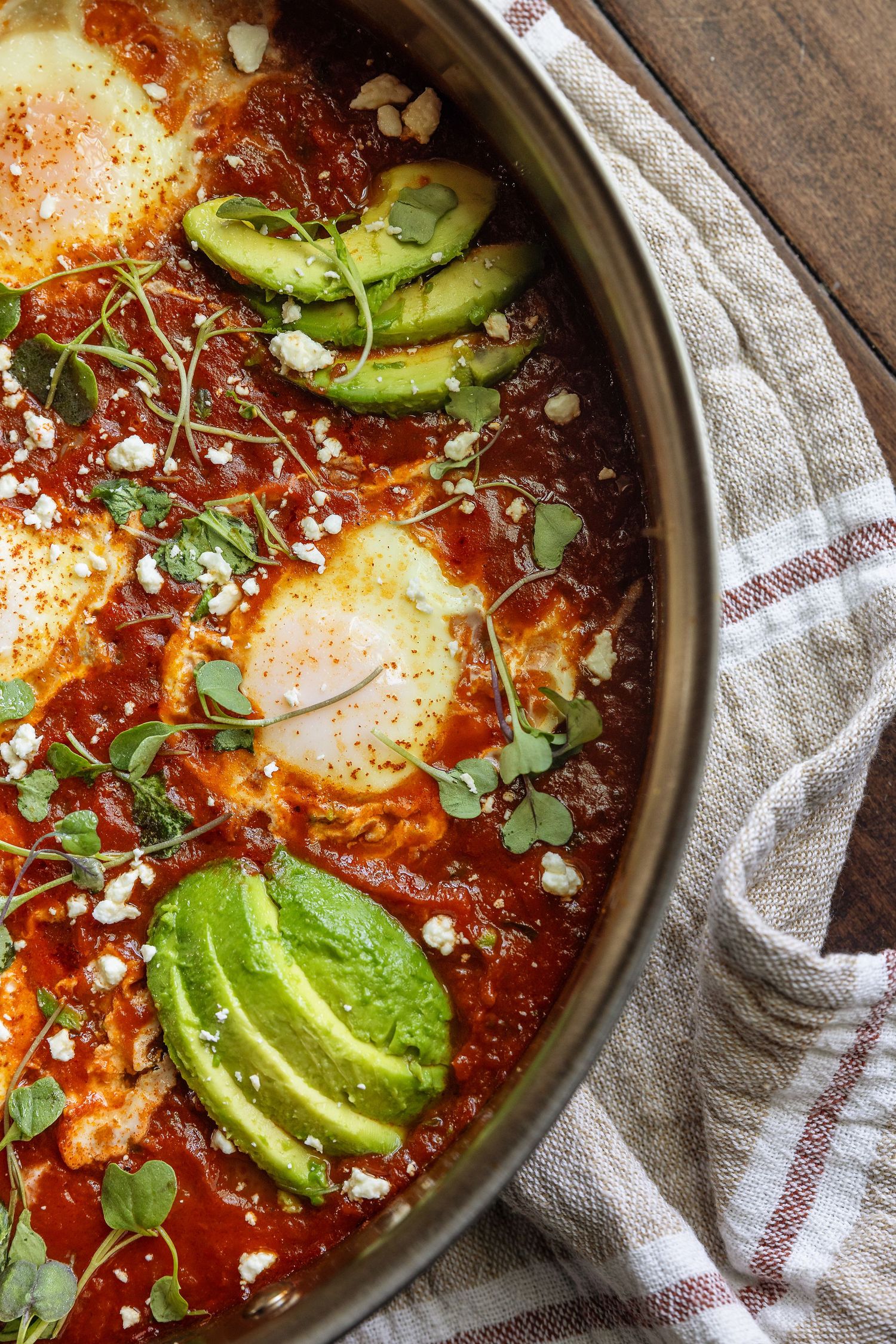 Shakshuka with poached eggs, sliced avocado, feta cheese, and fresh herbs in a tomato sauce skillet.