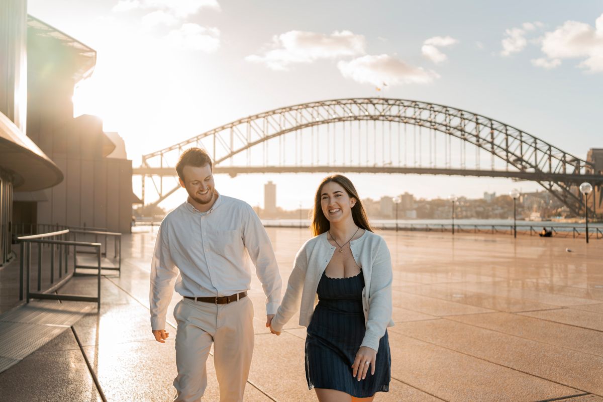 couple walking towards the photographer behind the Sydney opera house with a nice view on the harbour bridge couple photoshoot