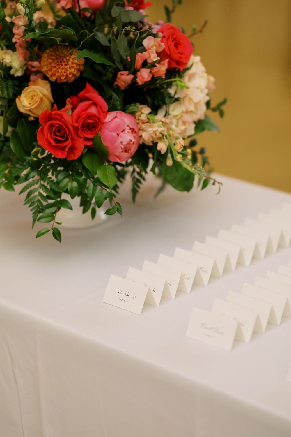 Wedding place cards arranged in neat rows on white tablecloth with colorful floral centerpiece of roses and greenery at Hall of Springs.