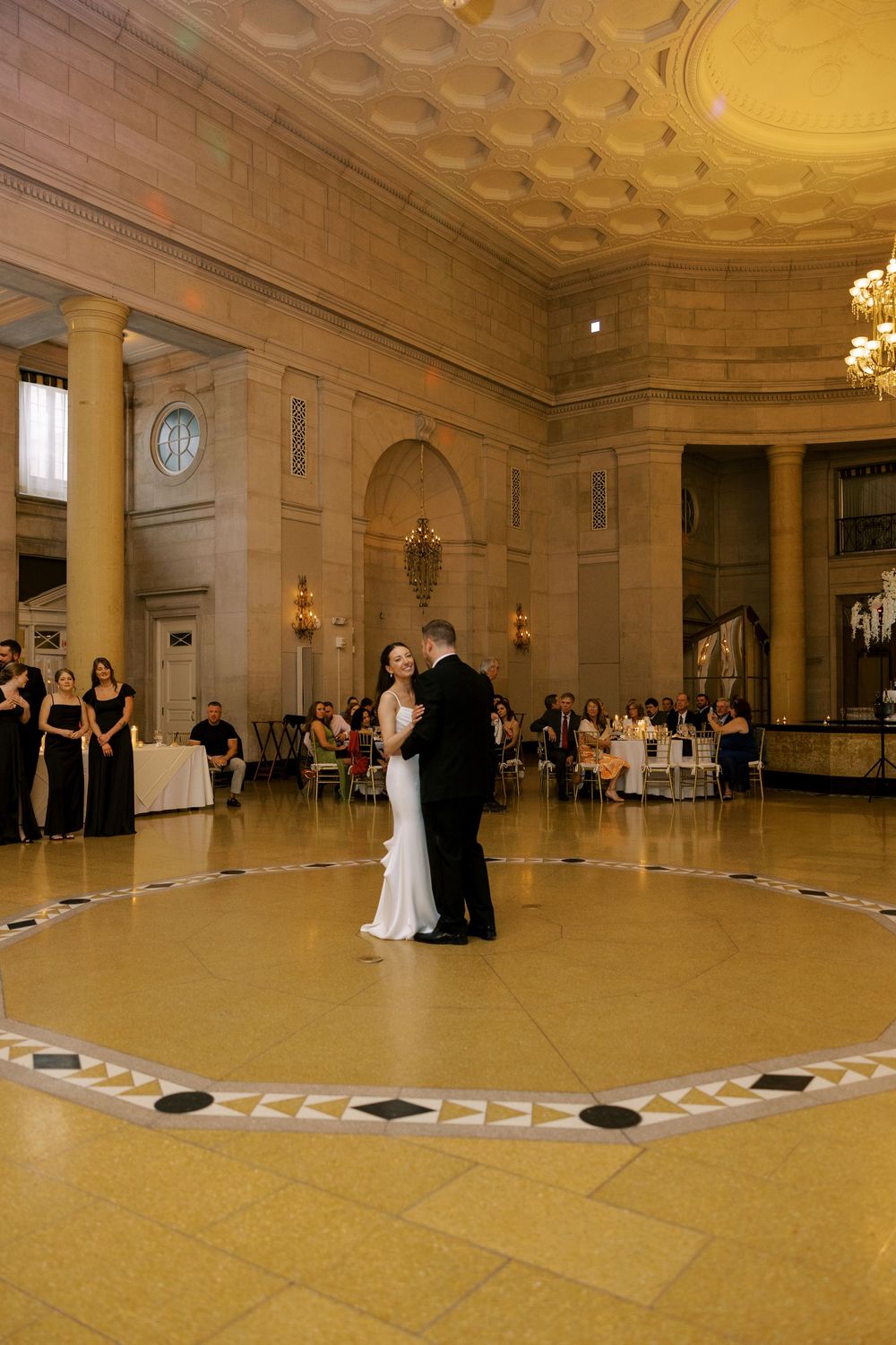 A couple shares their first dance in an elegant ballroom with ornate ceiling and columns while guests watch from the sides at Hall of Spring