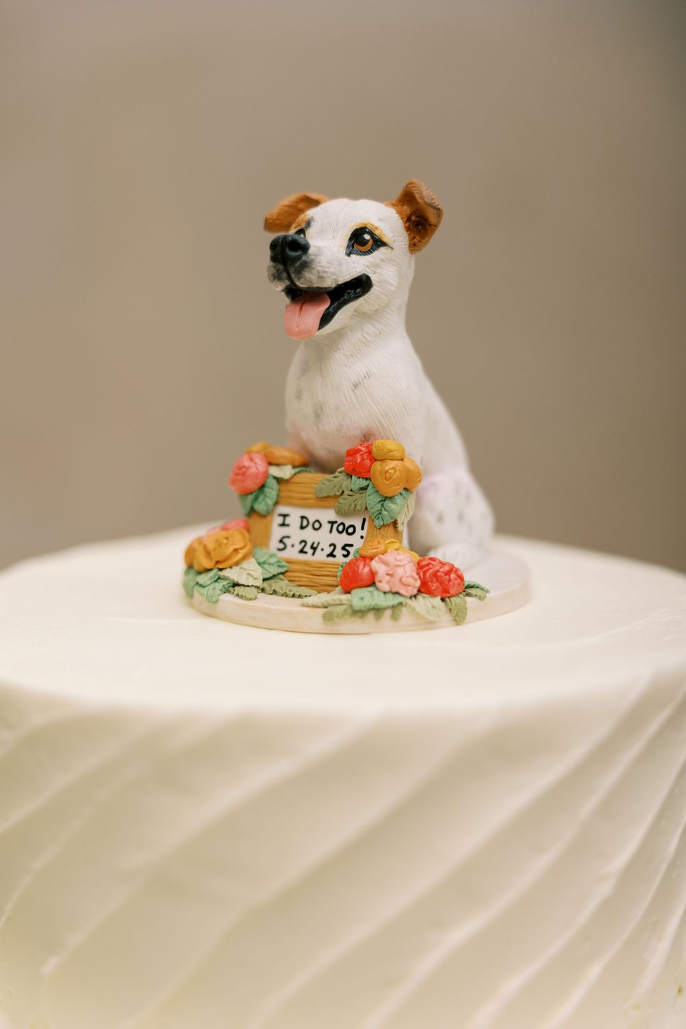 A smiling Jack Russell Terrier wedding cake topper adorned with orange flowers and a small sign on a white surface at Hall of Springs.