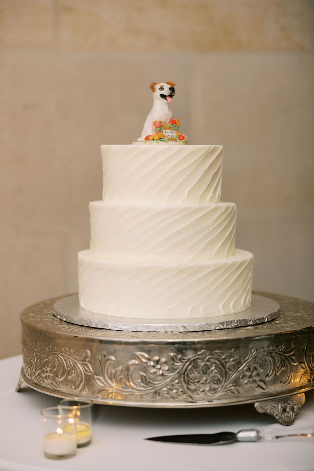 Three-tiered white wedding cake with diagonal textured frosting on an ornate silver stand with votive candles nearby at Hall of Springs.
