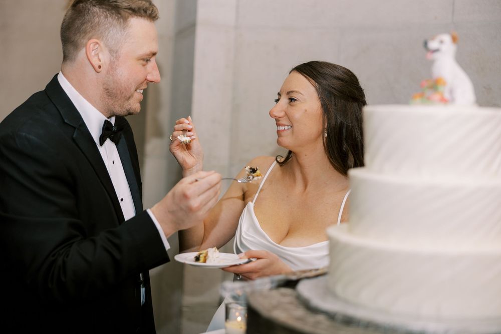 A couple in formal wedding attire share a playful moment while eating cake next to a white tiered wedding cake at Hall of Springs.
