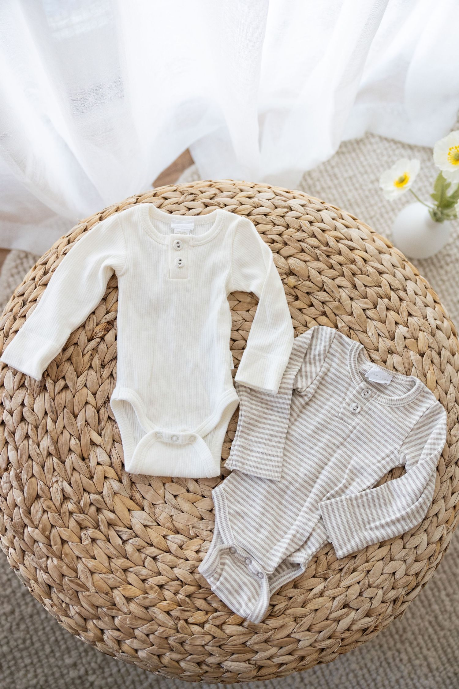 Two baby onesies laid on a round woven straw mat with white flowers, one solid white and one striped beige design.