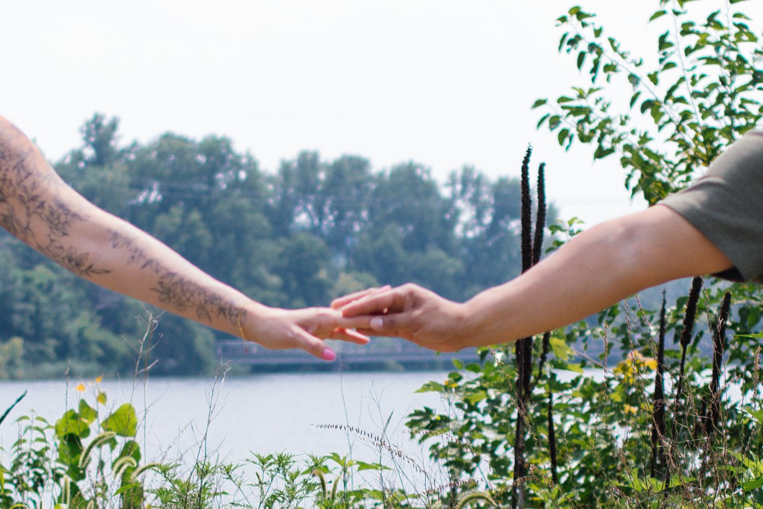 Couple holding hands against scenic lake backdrop with lush green trees in the background.