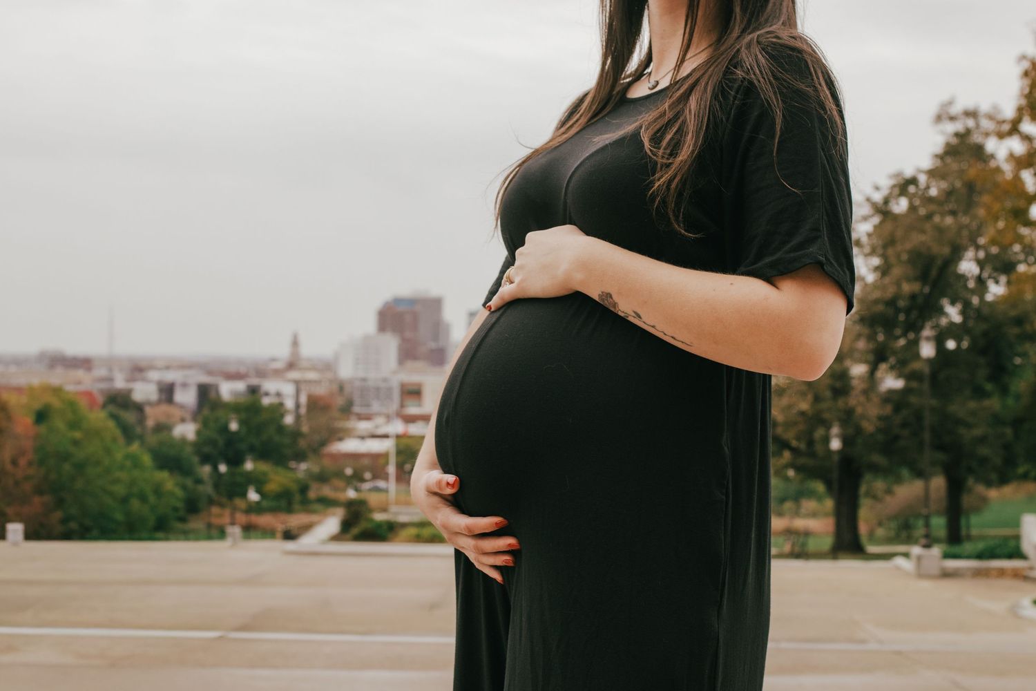 Pregnant woman in black dress cradling baby bump outdoors with city skyline in background.