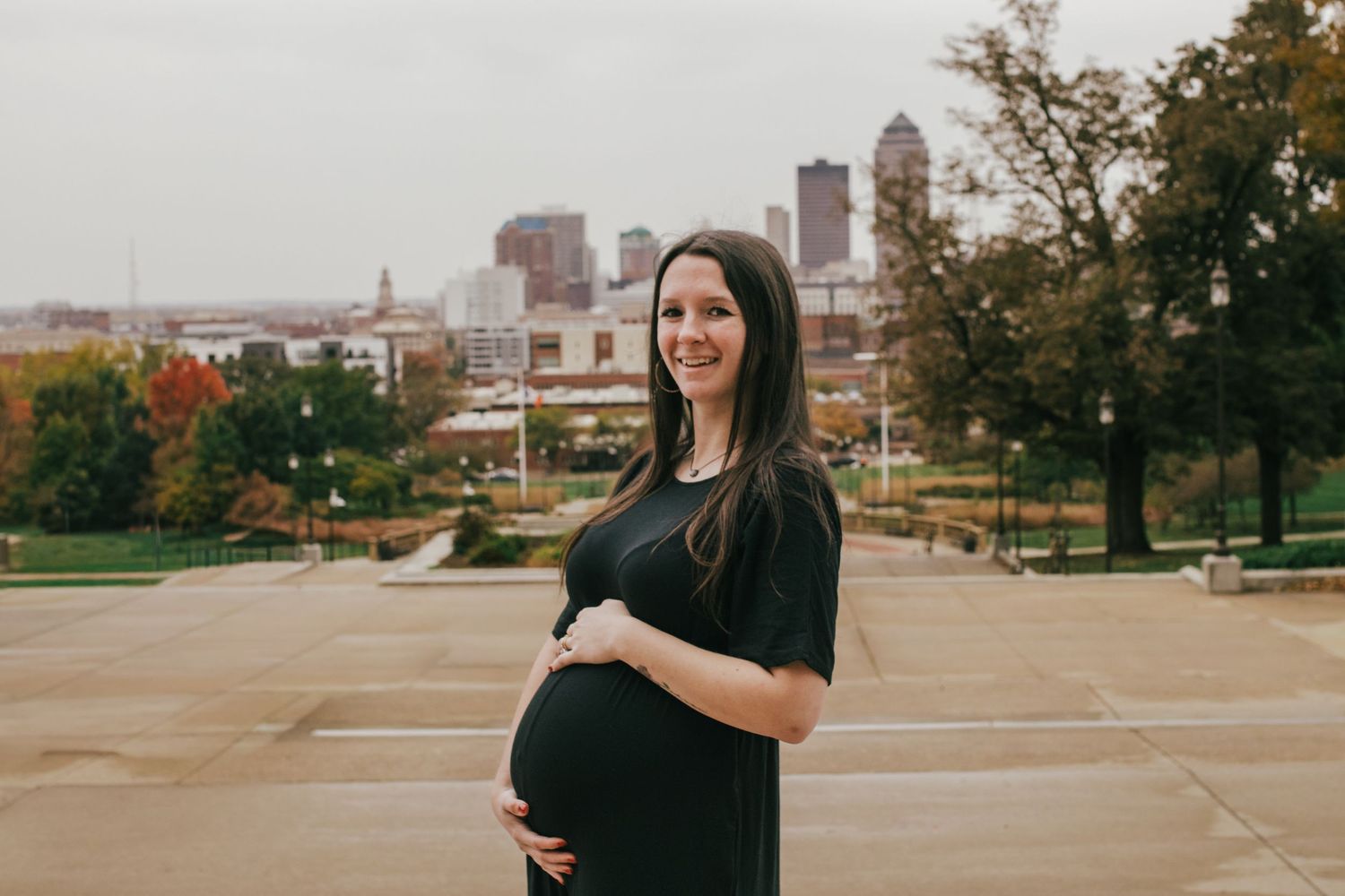 Smiling pregnant woman in black dress cradling baby bump outdoors with city skyline backdrop on overcast day.