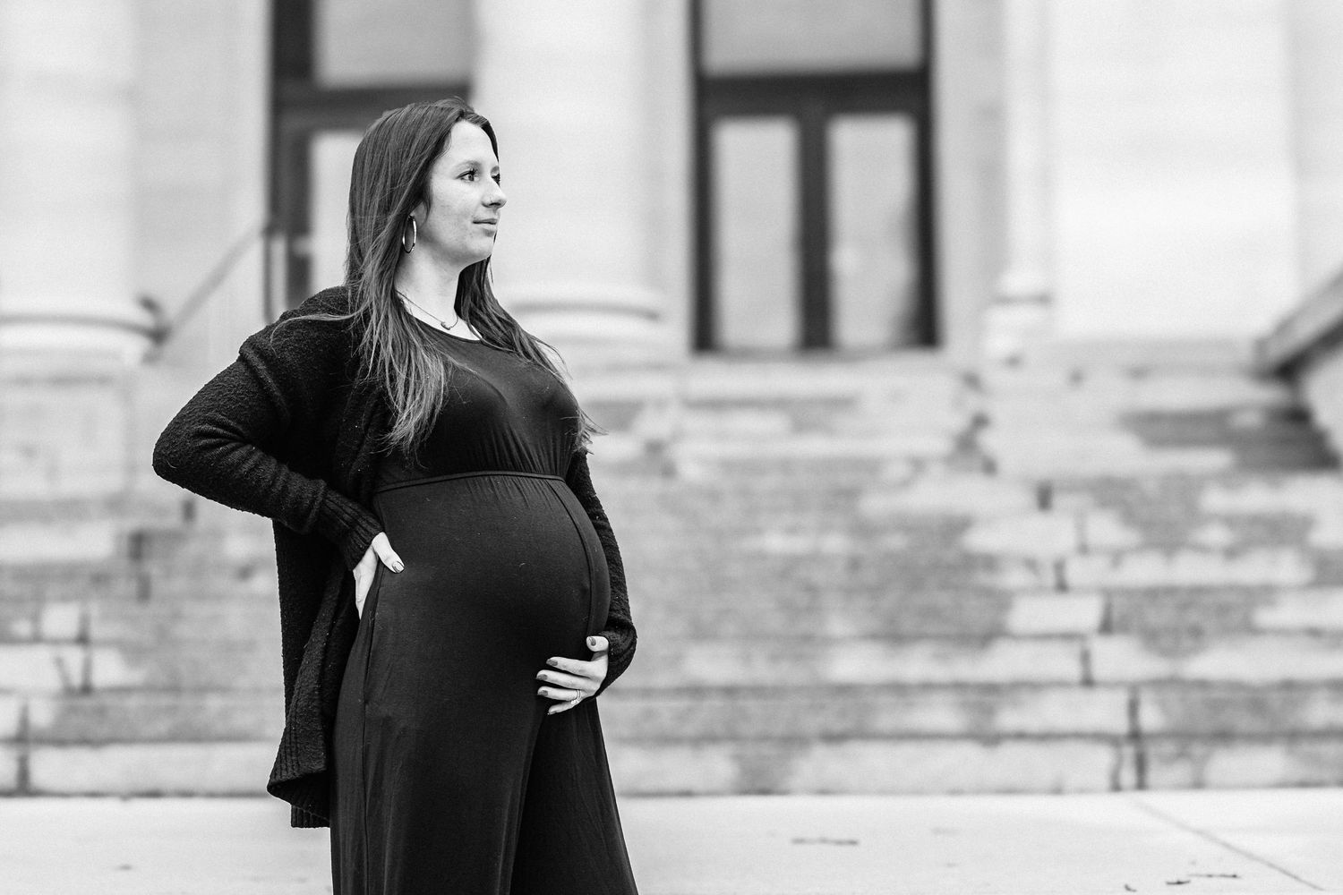 Black and white photo of a pregnant woman in a black dress cradling her baby bump outdoors on steps.