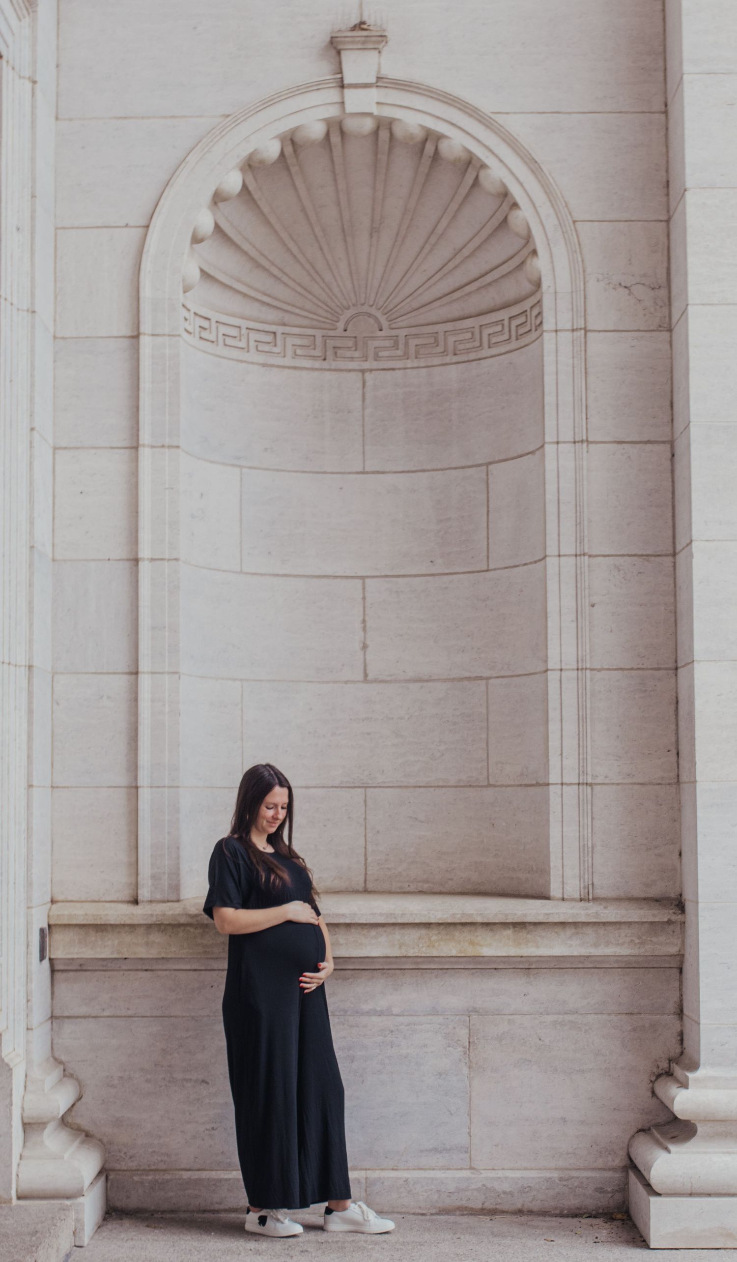 Pregnant woman in black maxi dress cradling baby bump, standing in ornate stone archway niche.