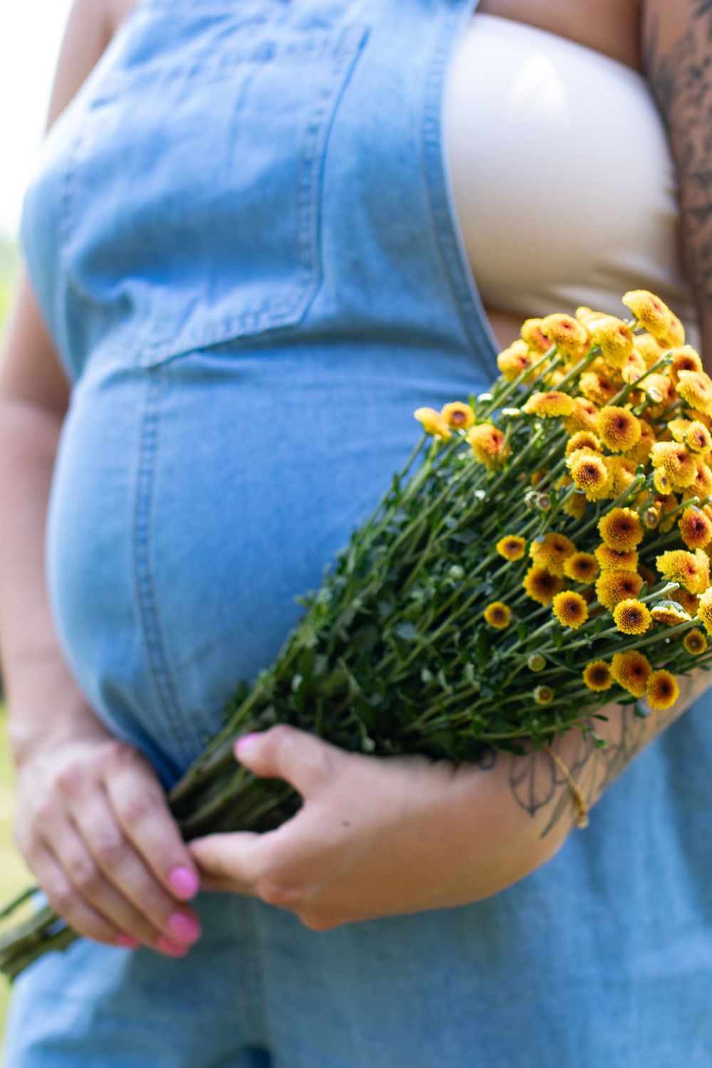 Person in denim overalls holding a bouquet of bright yellow flowers against a sunny outdoor background.