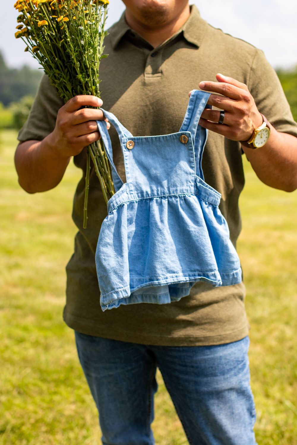 Someone in olive green holding flowers and denim overalls in a sunny field.