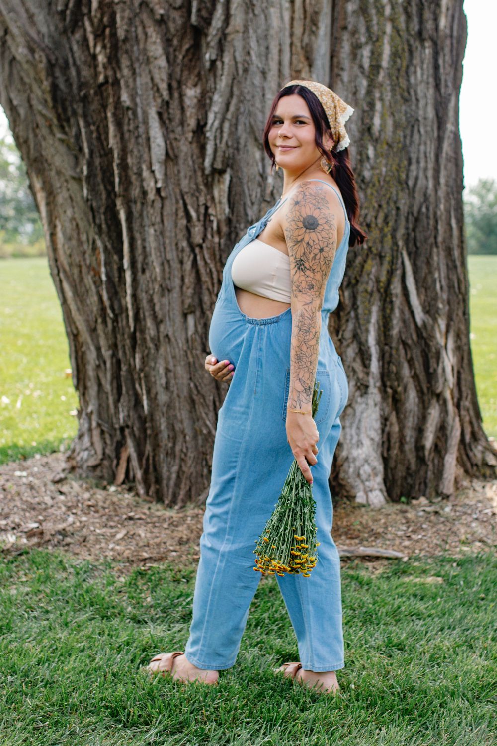 Person in denim overalls poses playfully near large tree trunks in a grassy field.