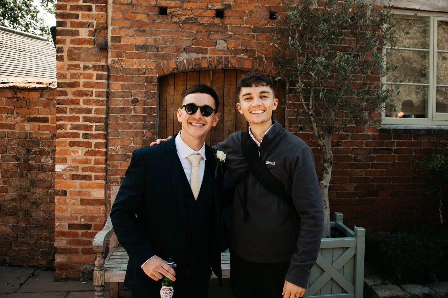 Two friends stand together smiling in front of a red brick wall wearing sunglasses and casual outerwear.