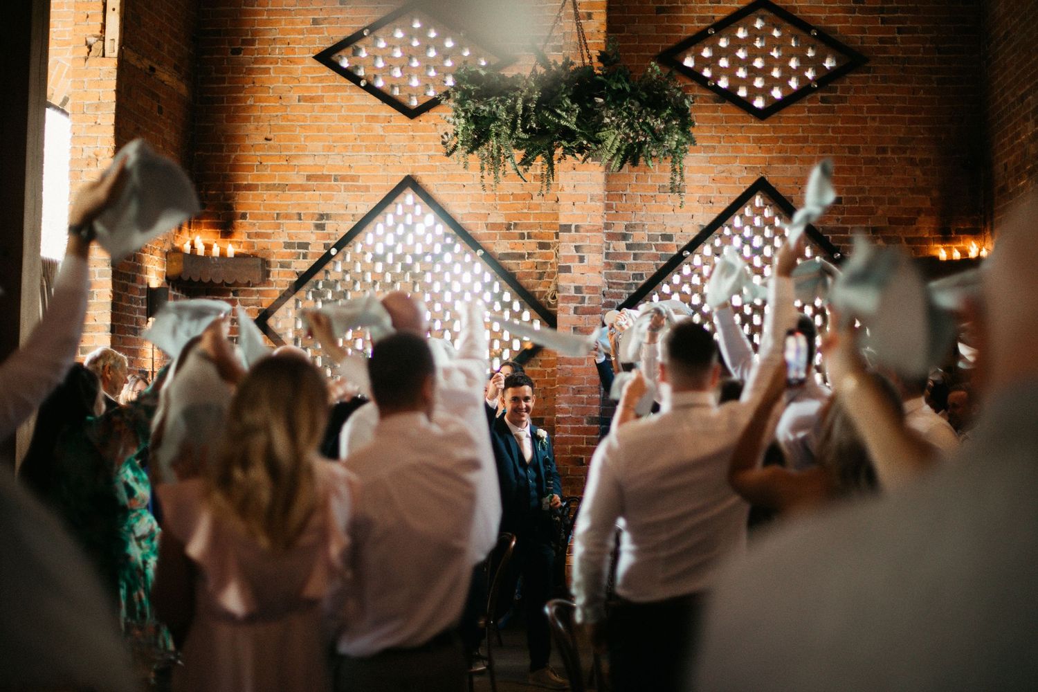 A couple stands in elegant wedding attire beneath a rustic brick archway with wooden doors at a historical venue.