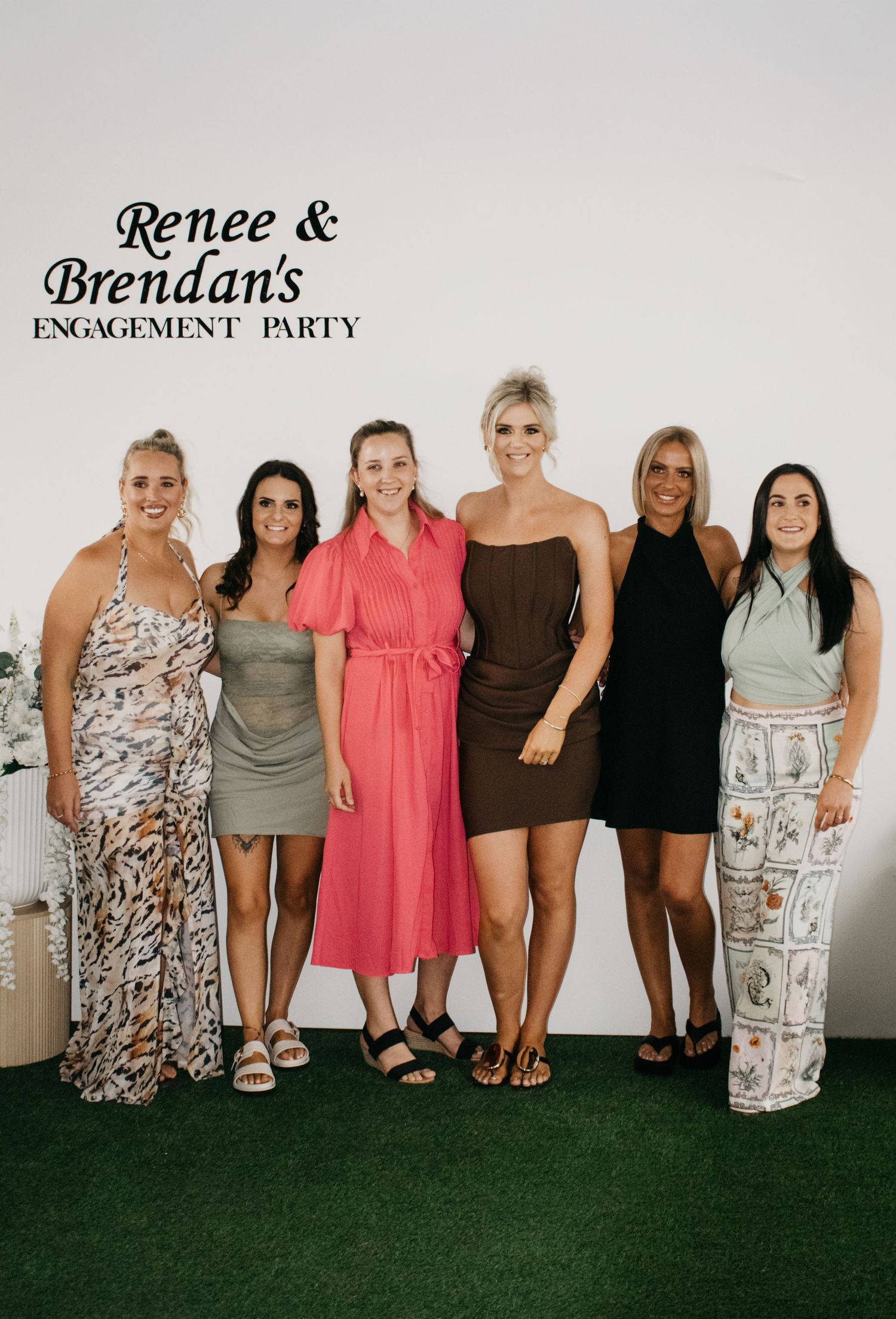 A group of people stand together in a row wearing various outfits including dresses and swimwear.