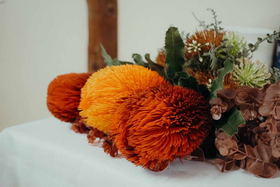 Orange Banksia flowers with dried foliage arranged on white linen surface in natural lighting.
