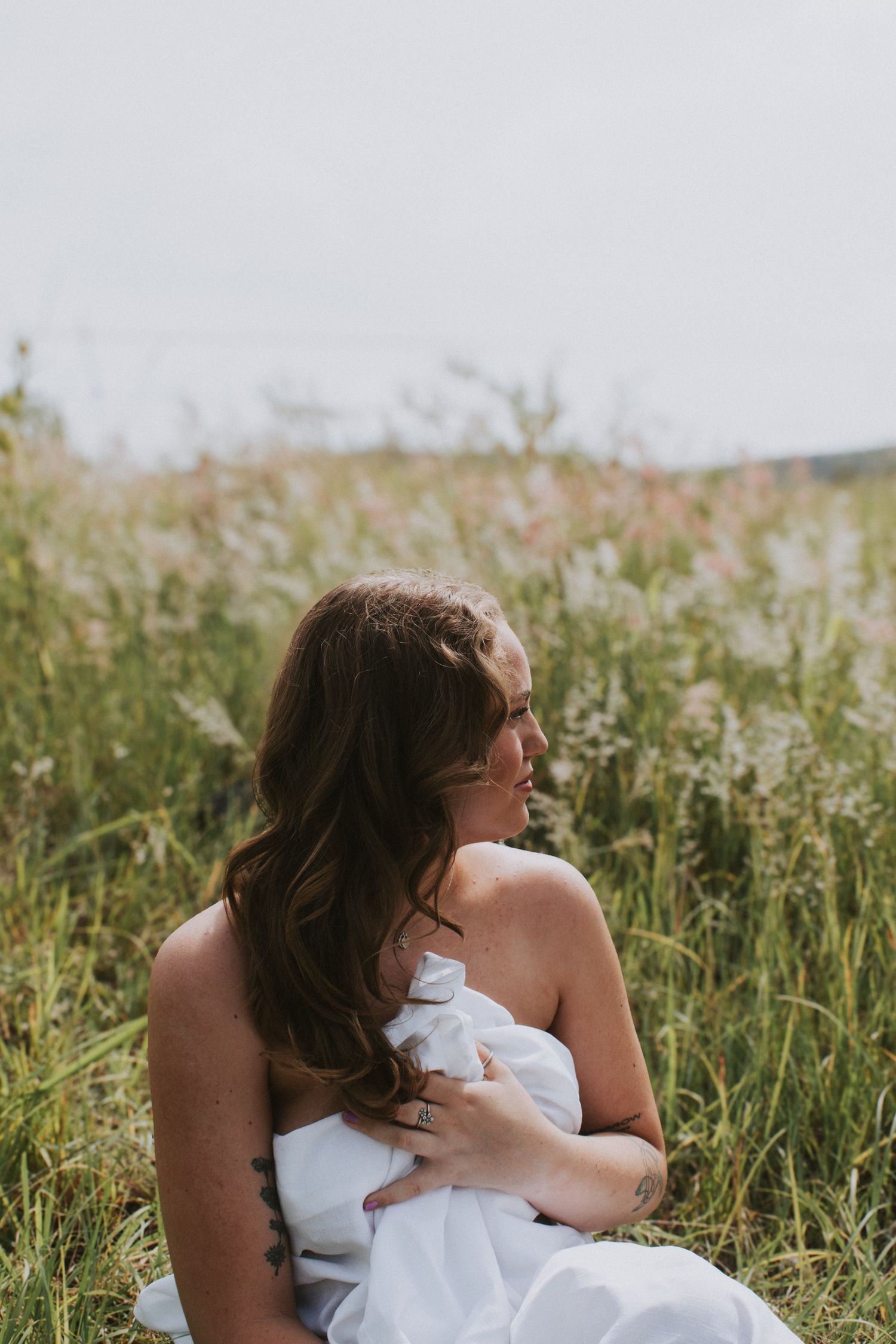 Woman in white dress sitting in tall grass meadow during golden hour looking to the side.