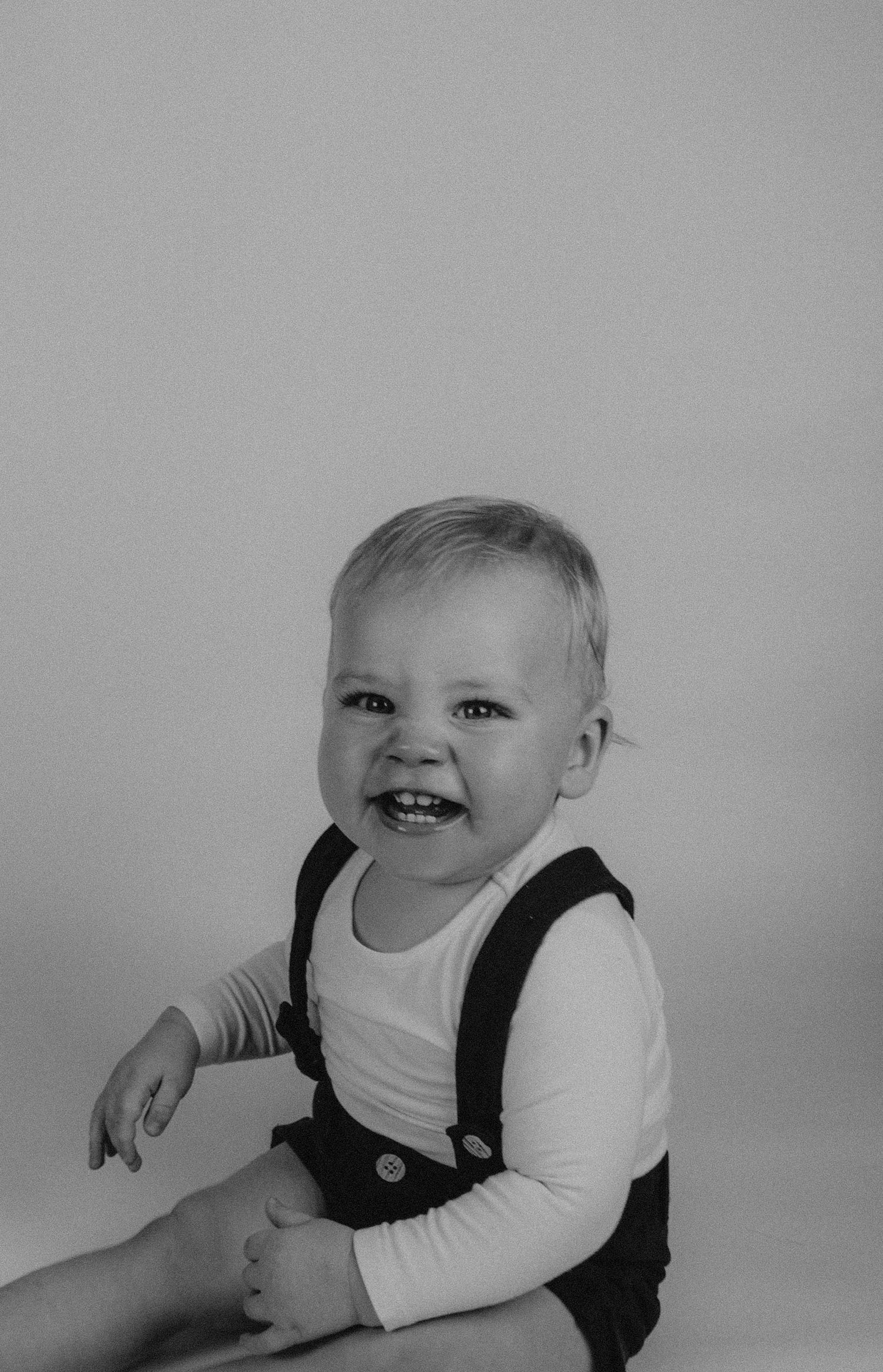 Black and white portrait of a smiling toddler wearing overalls and a white long sleeve shirt against a plain background.