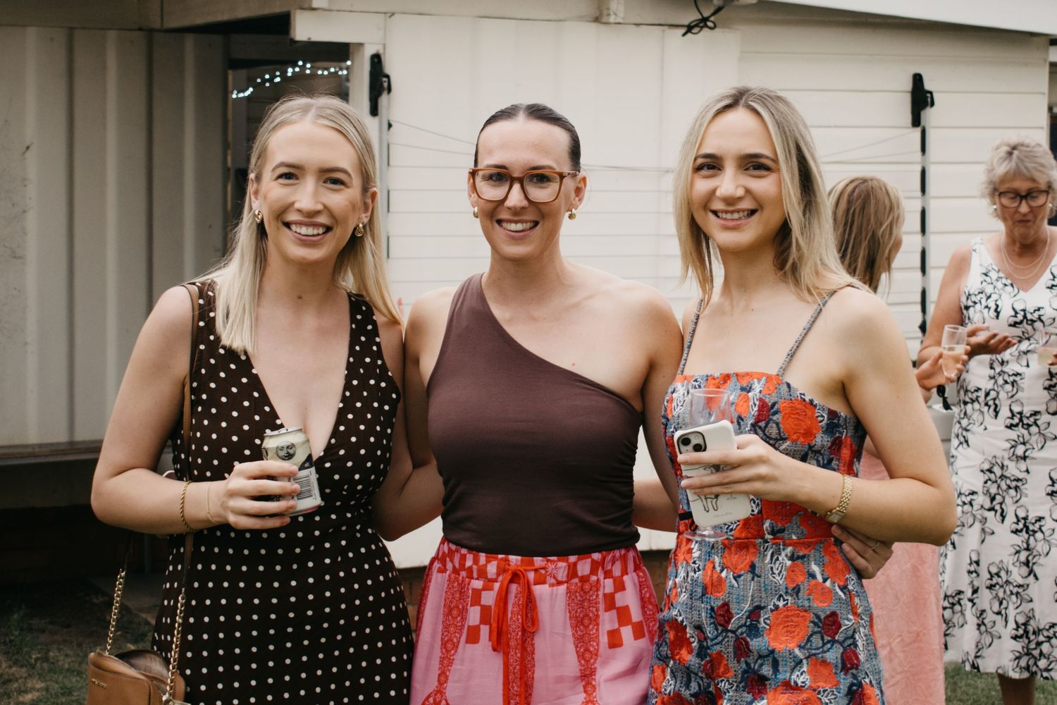 A group of friends in summer dresses stand together smiling at an outdoor event.