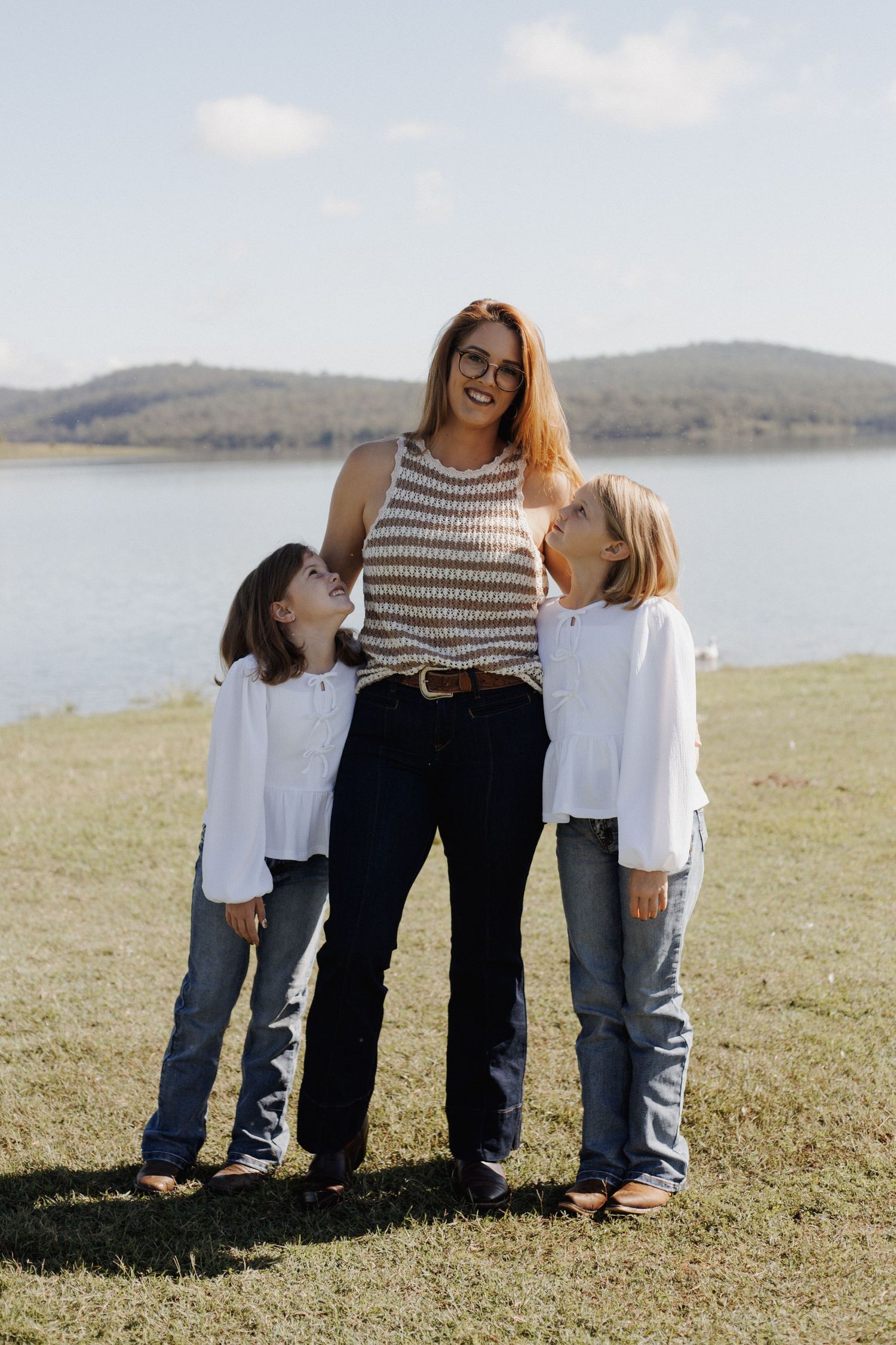 A caring figure in a striped tank top stands with two children by a scenic lake on a grassy hillside.