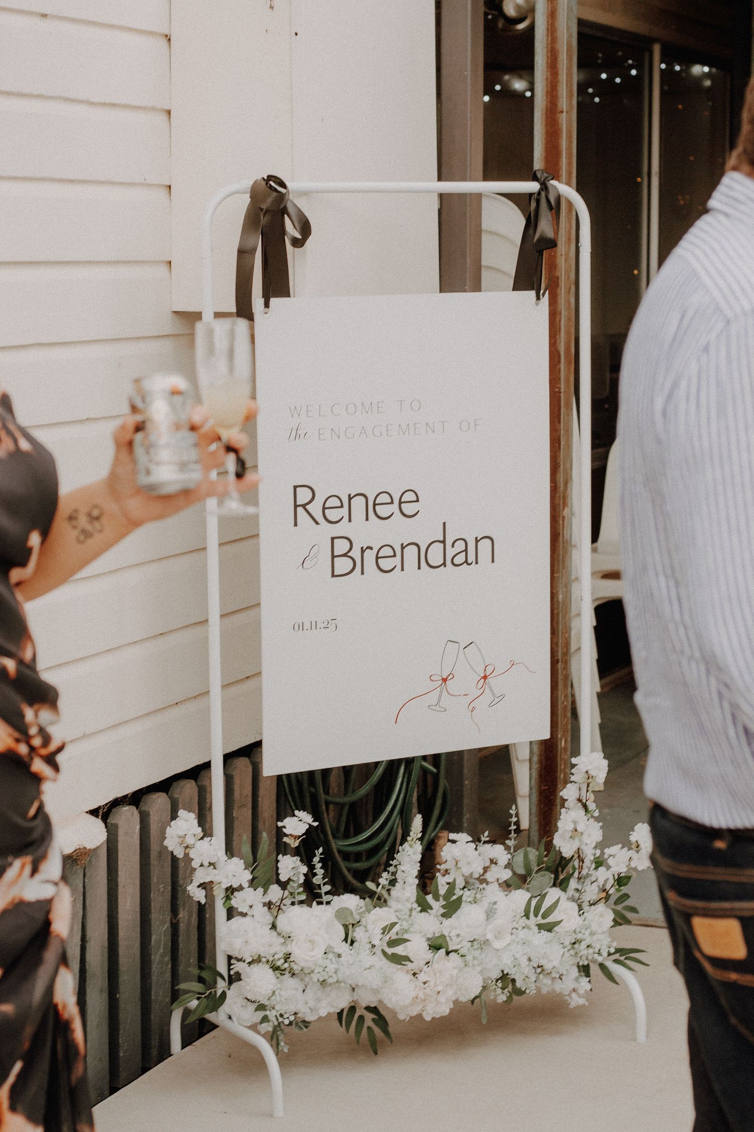 Wedding reception signage with floral decorations displays elegant name cards against a white wall.