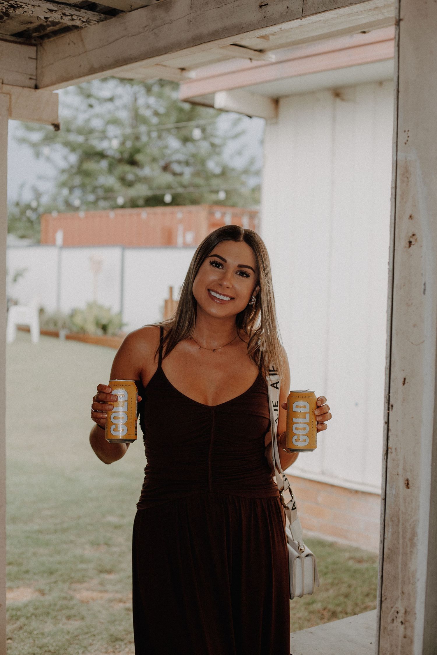 Three women in black dresses hold up beverage cans while posing together on an outdoor porch.