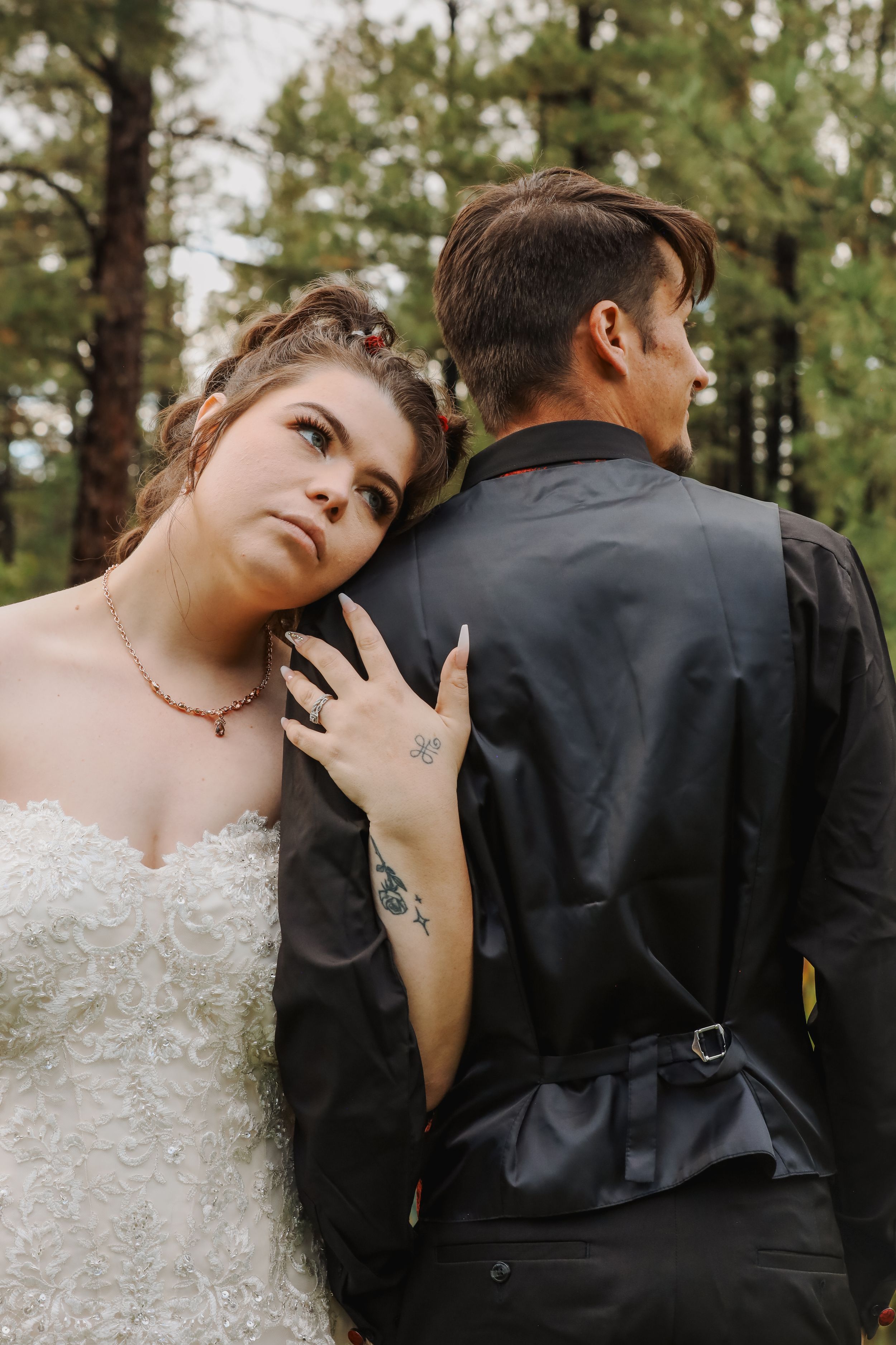 Bride in white strapless gown leans on groom's back in black suit among pine trees.