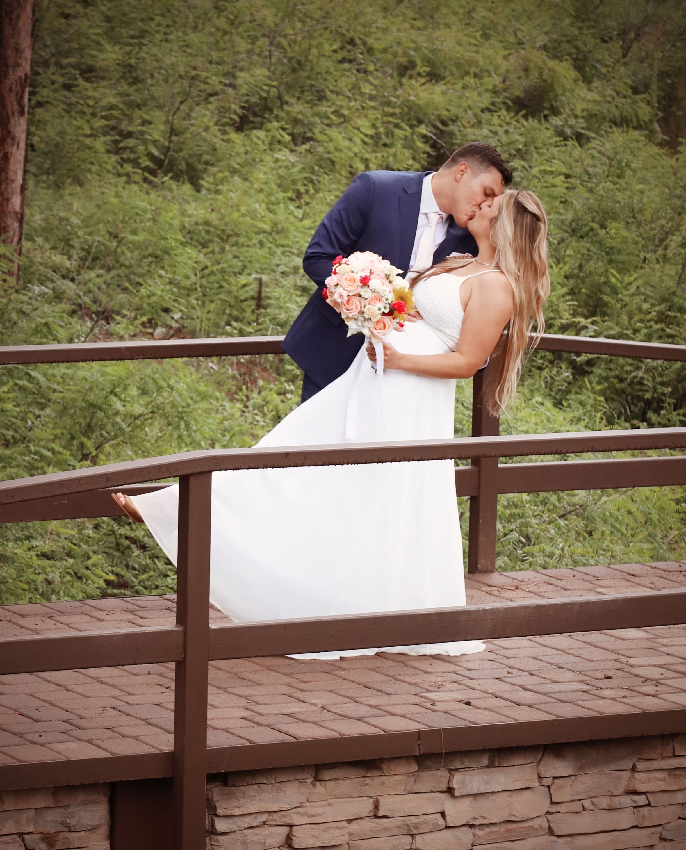 Bride and groom share a romantic kiss on a wooden bridge surrounded by lush green forest on their wedding day.