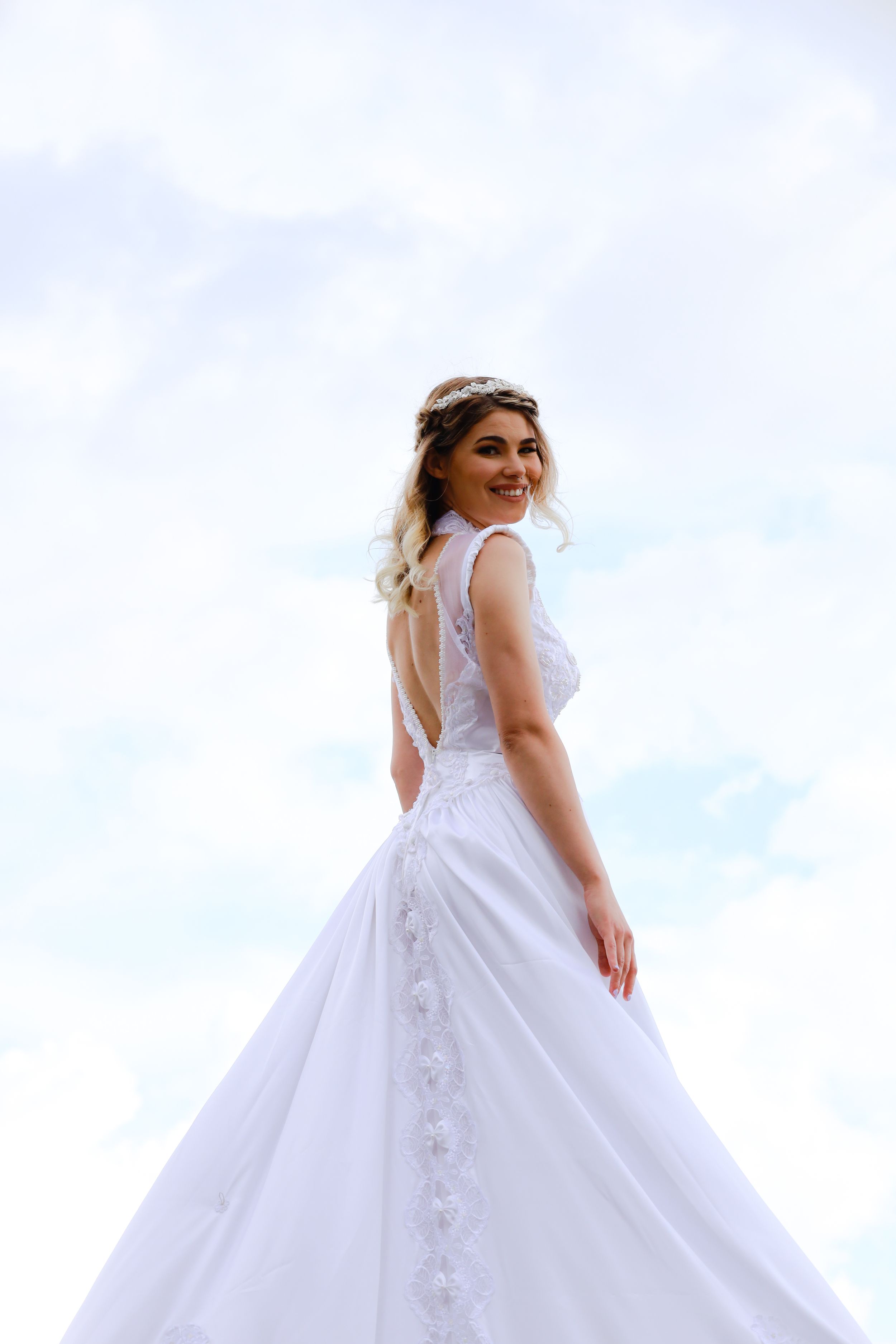 Beautiful bride in white lace backless wedding gown with floral crown smiling against a cloudy sky.