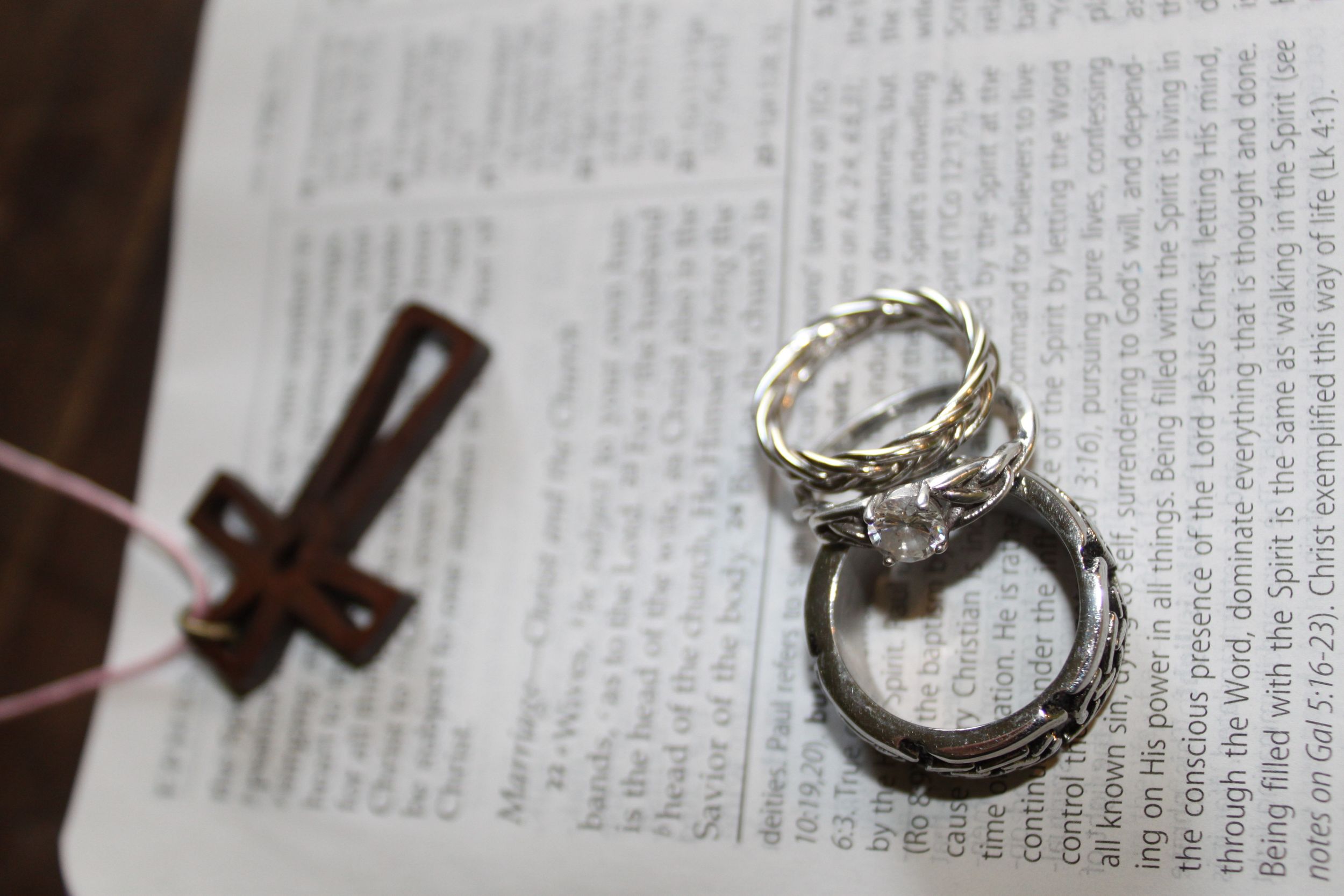 Two silver wedding rings rest on an open Bible next to a wooden cross bookmark.