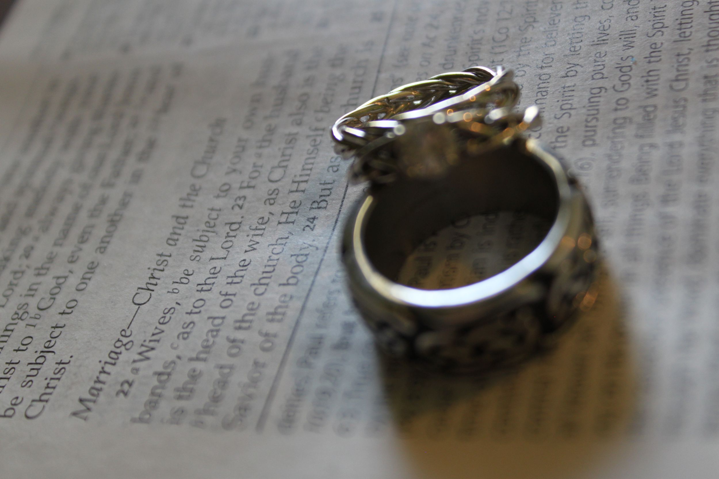 Two ornate silver wedding rings rest on an open Bible page in soft focus lighting.