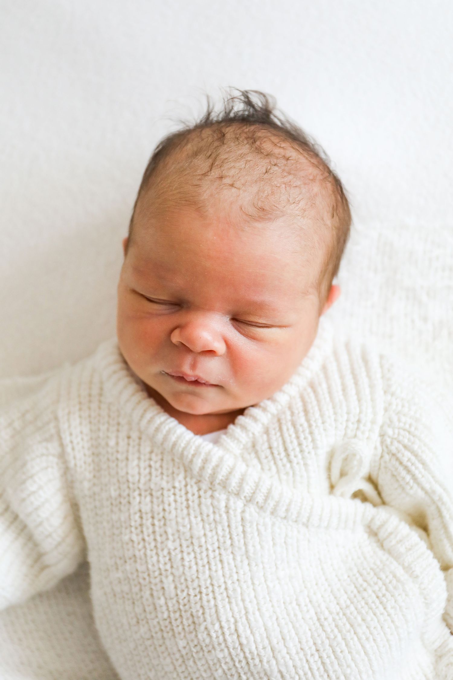 A peaceful sleeping newborn baby wrapped in a cozy white knit romper against a light background, during newborn shoot in Nottingham