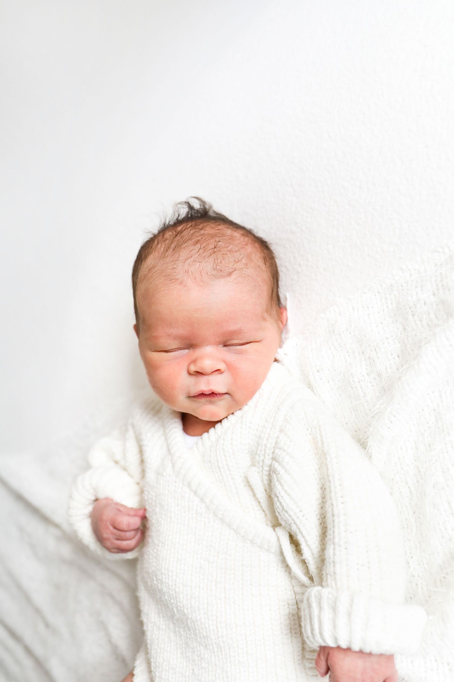 A peacefully sleeping newborn baby wearing a soft white knit romper lies on white bedding, during newborn shoot in Nottingham