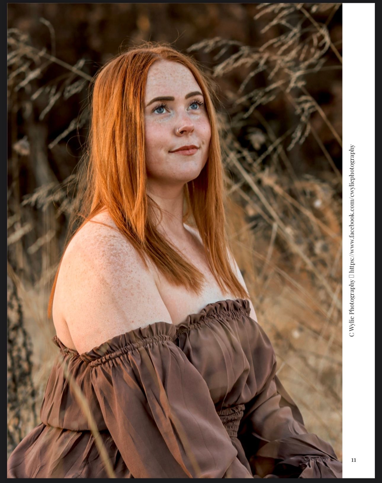 A woman with long red hair poses outdoors in a brown off-shoulder dress against a natural backdrop at sunset.