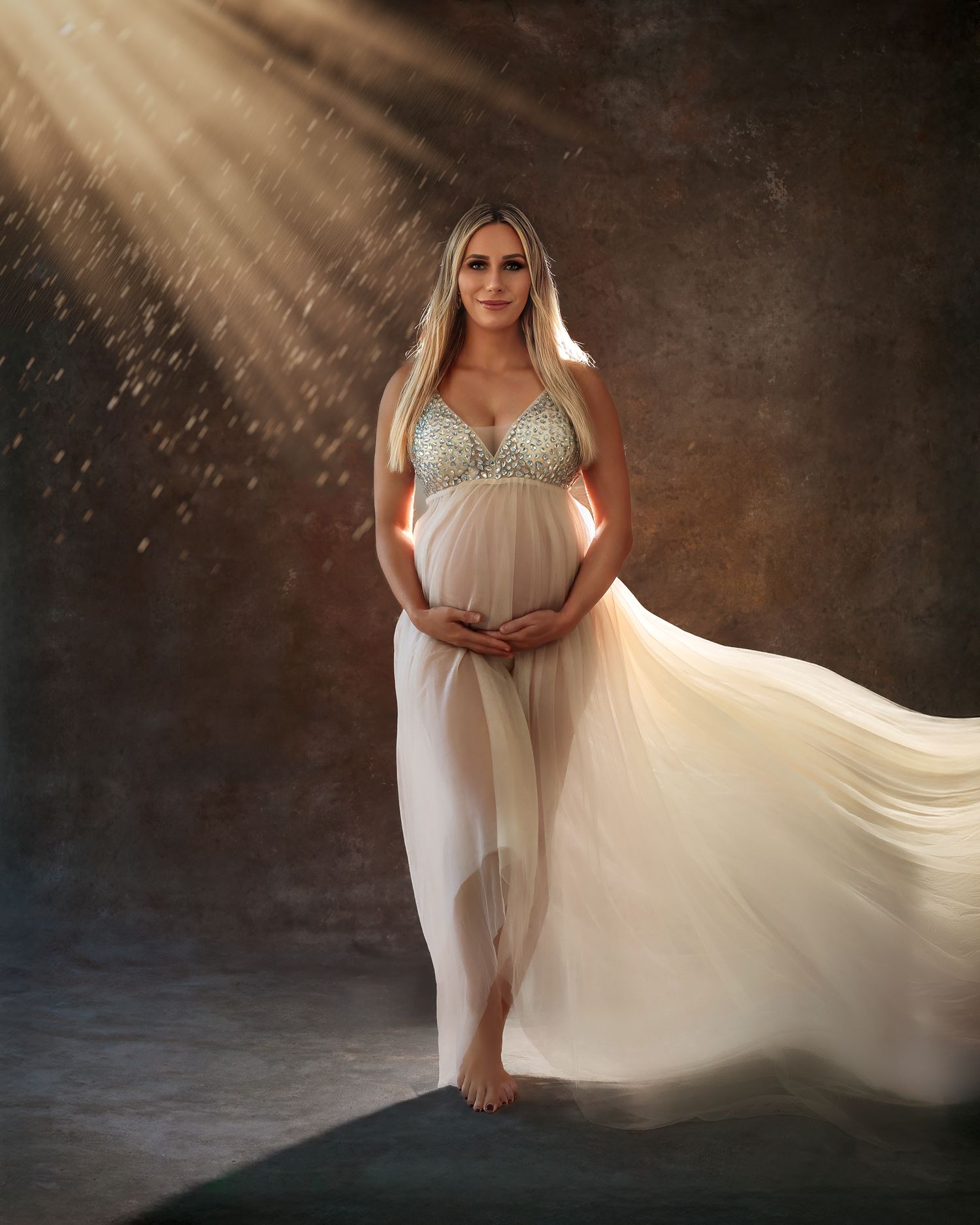 Dreamy studio portrait: woman in a cream flowing maternity gown with jewel embellishments on the bust, stands beneath ethereal light beams.