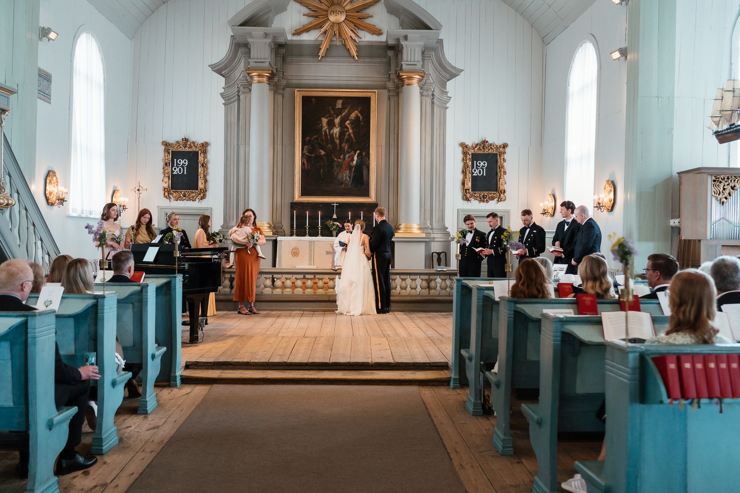 Traditional church wedding ceremony taking place in a bright white chapel with teal-colored pews and ornate golden altar.