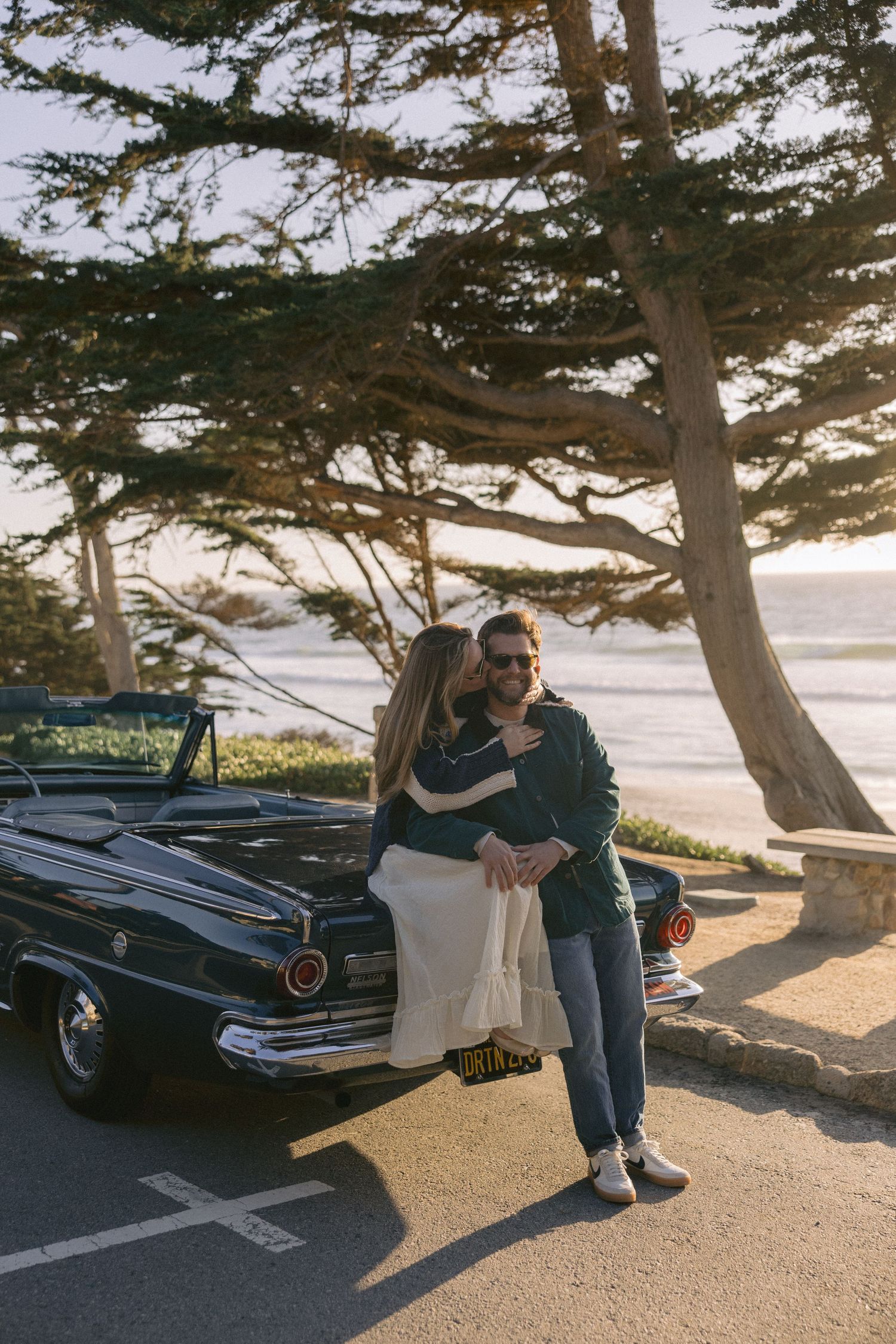 A couple embraces by a vintage black car parked beneath cypress trees along a coastal roadway at sunset.