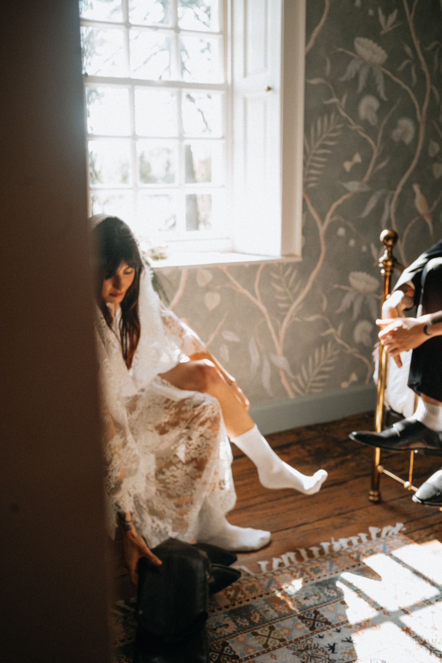 Bride in lace wedding dress and white boots sits by sunlit window with floral wallpaper on wooden floor.