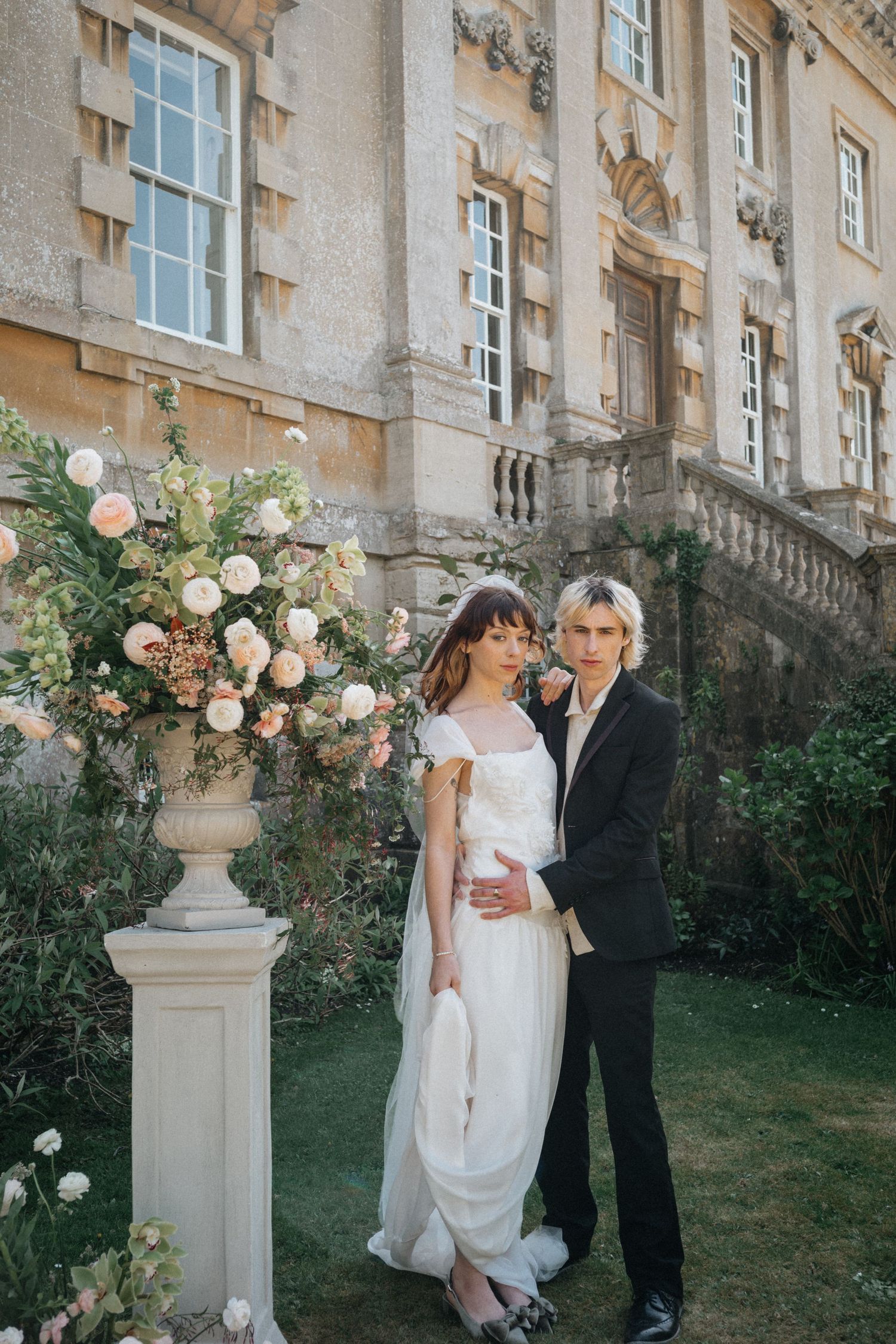 Bride in white gown and groom in black suit pose by floral urn outside a grand stone manor.