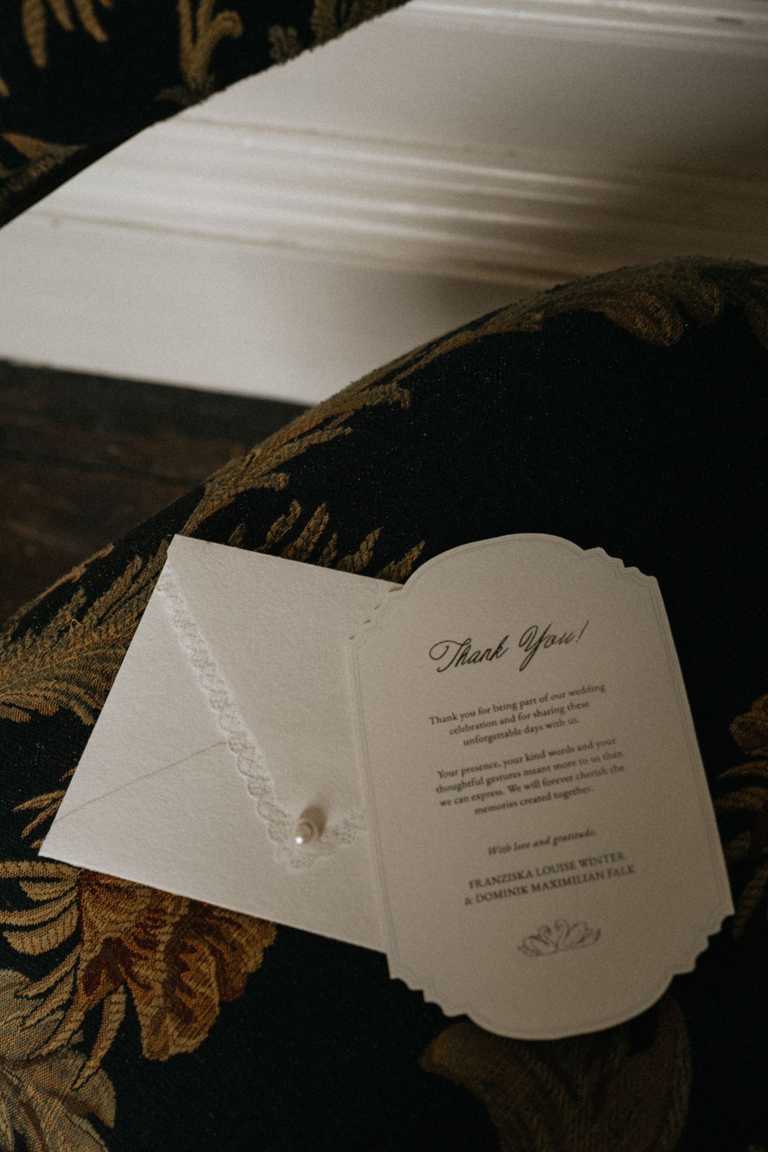 Elegant wedding thank you card with lace envelope on dark floral velvet chair.