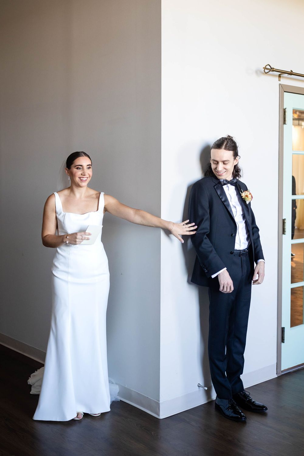 A bride in a white gown and groom in a dark suit pose playfully with a wall between them before their first look.