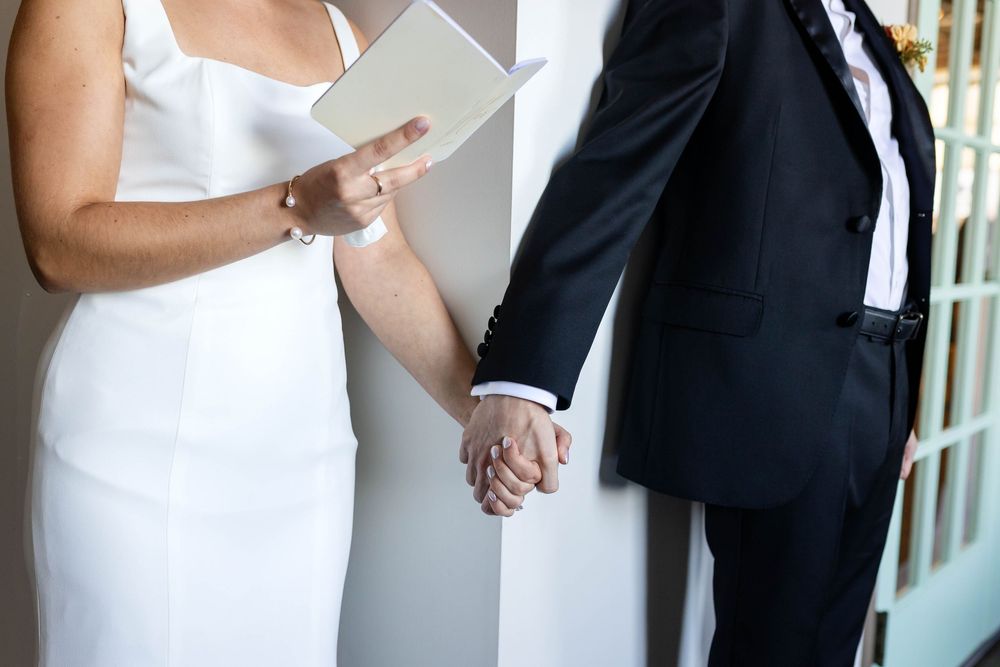 Bride and groom hold hands while reading wedding vows, dressed in elegant white dress and black suit.