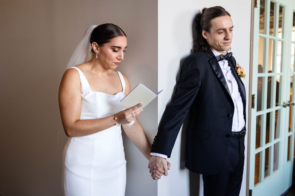 A bride reads a letter while holding hands with her groom around a corner during their first look moment.