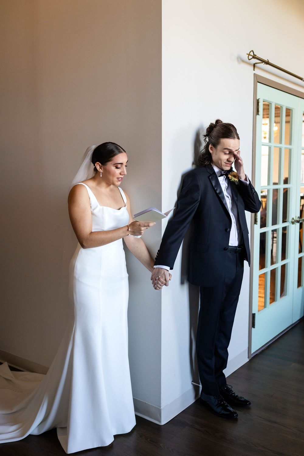 A couple stands together on their wedding day, holding hands while reading notes on opposite sides of a door.